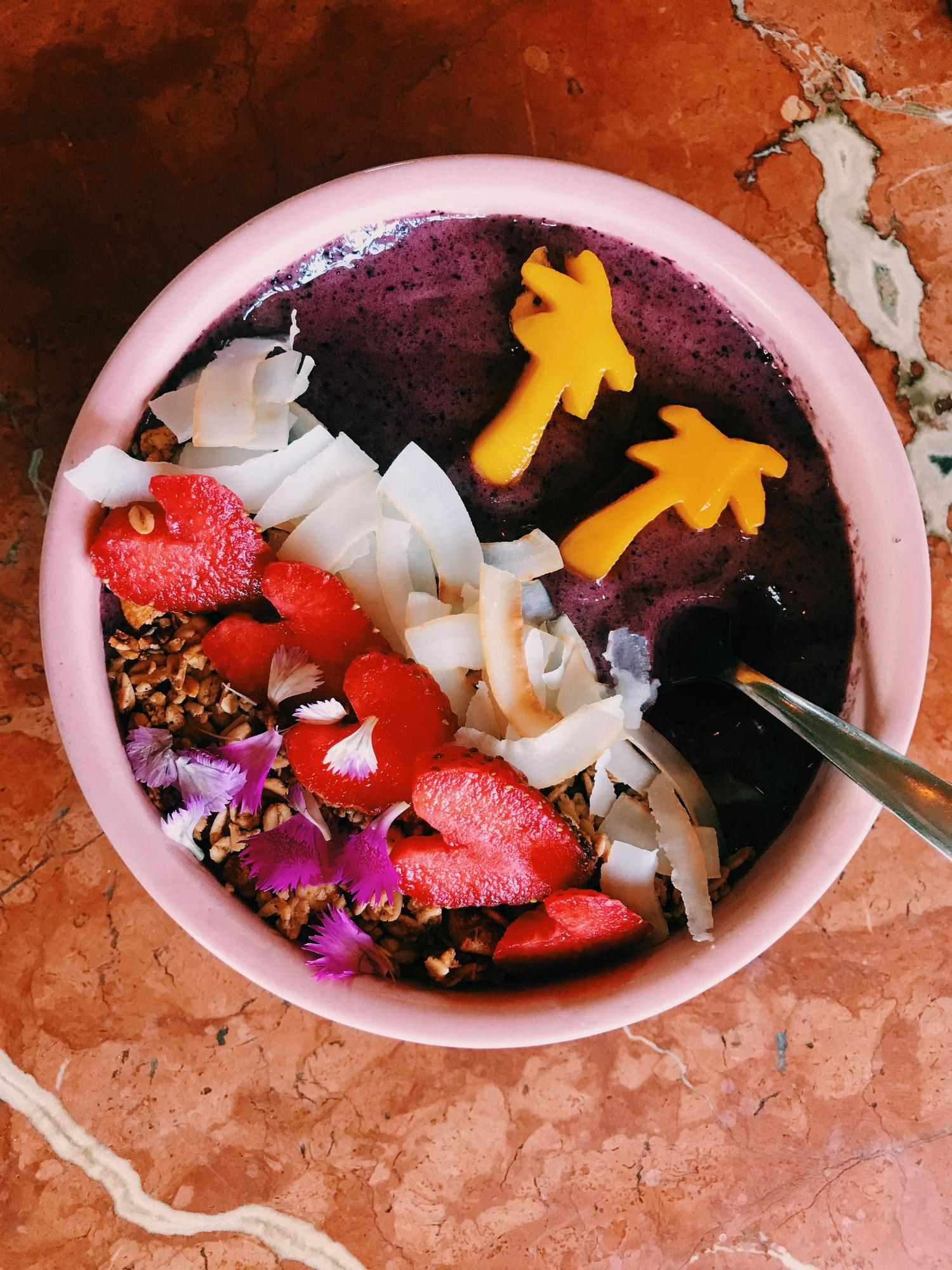 Colorful açaí bowl topped with granola, coconut flakes, mango palm trees, strawberries, and edible flowers – tropical superfood breakfast