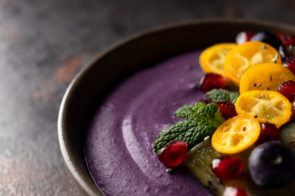 Belgian açaí gastronomy signature dish featuring purple açaí purée in artisanal ceramic bowl topped with kumquat slices, mint leaves and pomegranate seeds